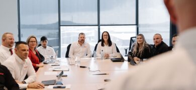 Join GoFox team members sitting around a boardroom table during a meeting, with a presenter speaking at the front and large windows in the background.
