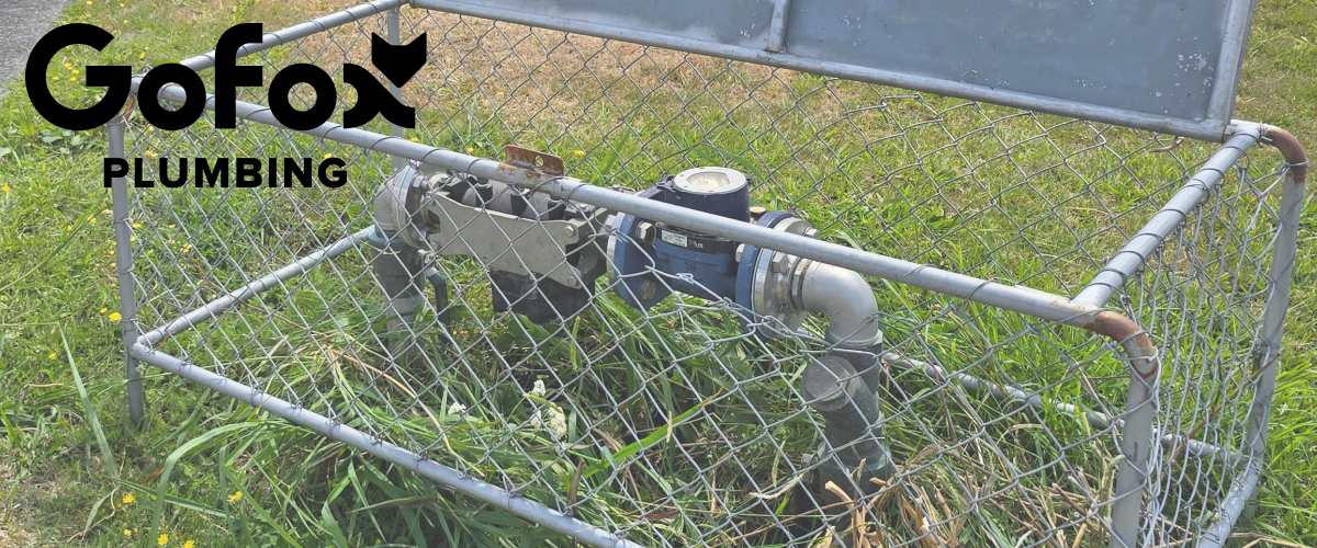 Backflow prevention testing device enclosed in a chain-link security cage at a commercial property in New Zealand, with GoFox Plumbing branding overlay.